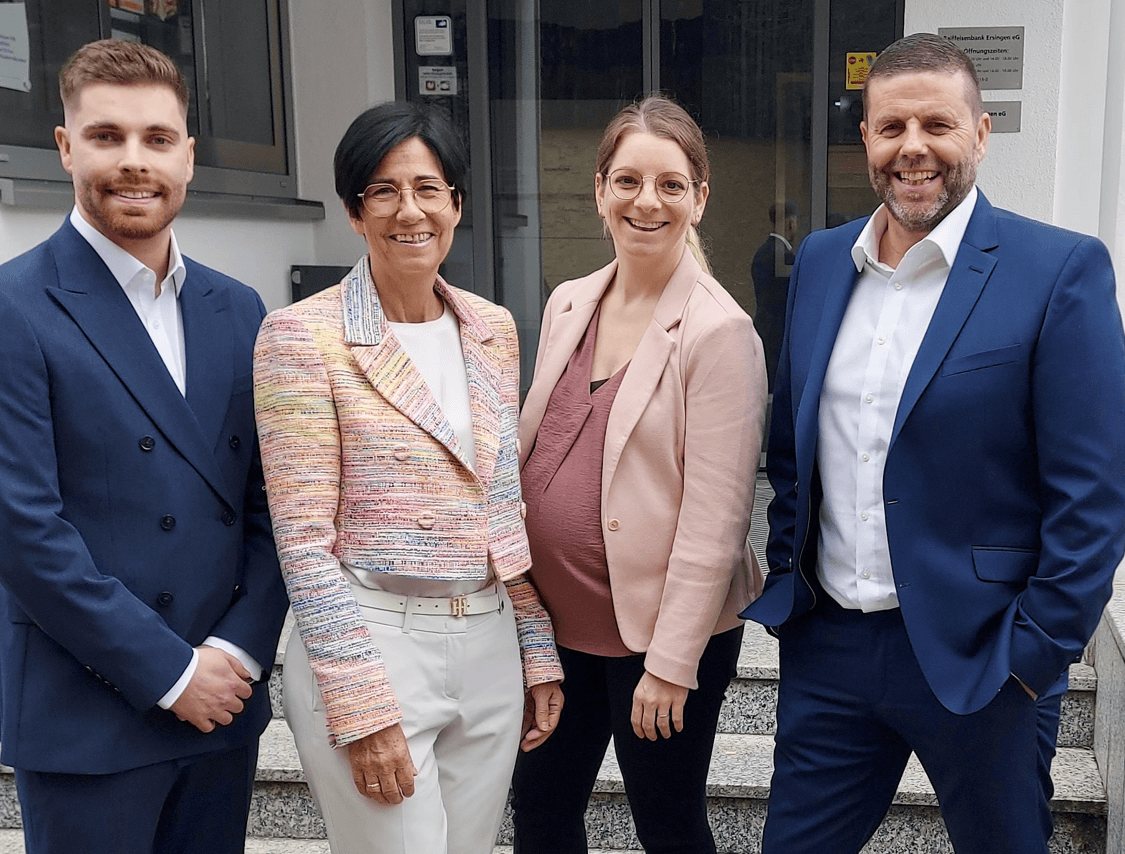 Vier lachende Personen in Businesskleidung stehen gemeinsam vor einem Gebäude und posieren als Gruppe für ein Foto