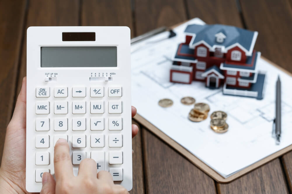 Eine Hand hält einen Taschenrechner, daneben liegen Münzen und ein kleines Hausmodell als Symbol für die Kosten beim Immobilienverkauf